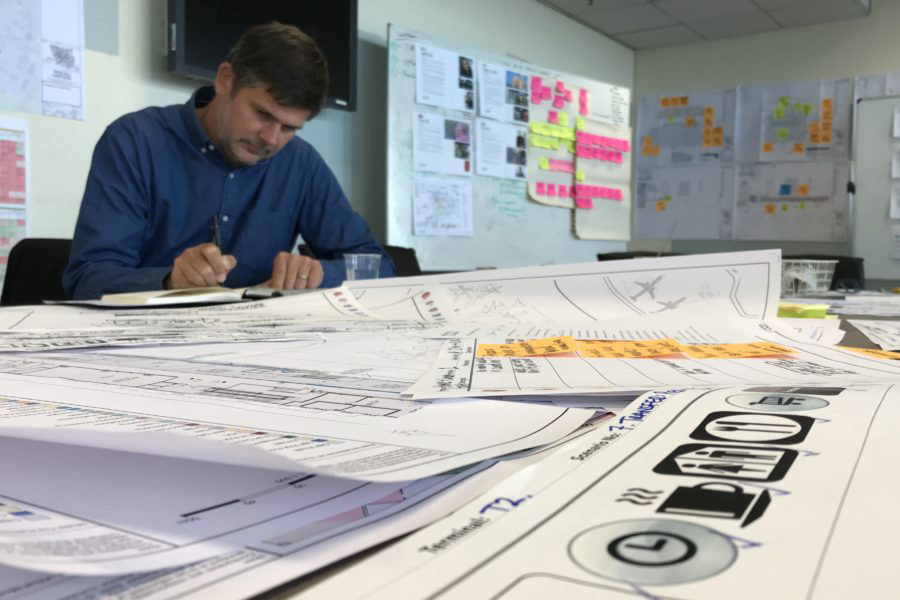 Phil, Head of Experience Design at Mima, is white and wears a blue shirt. He is working at a table that is full of working papers. Walls of colourful post-it notes are behind him.