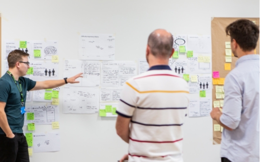 Three white males, all in casual clothing, face a whiteboard covered in paper and colourful post-it notes. They are in discussion, with one man presenting findings to the other two.